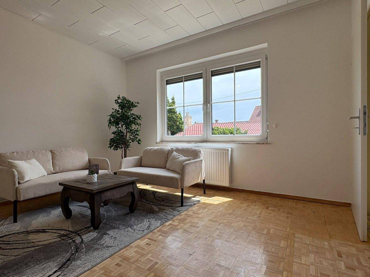 Helles Wohnzimmer mit Parkettboden, bequemen Sofas und großem Fenster mit Blick ins Grüne.