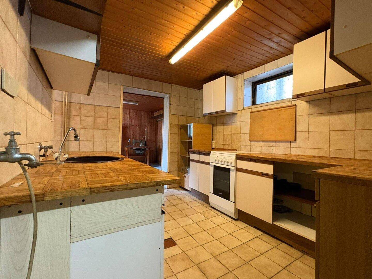 Einfache Küche im Untergeschoss mit Holzdecken, Fliesenboden und älteren Einbaumöbeln.
