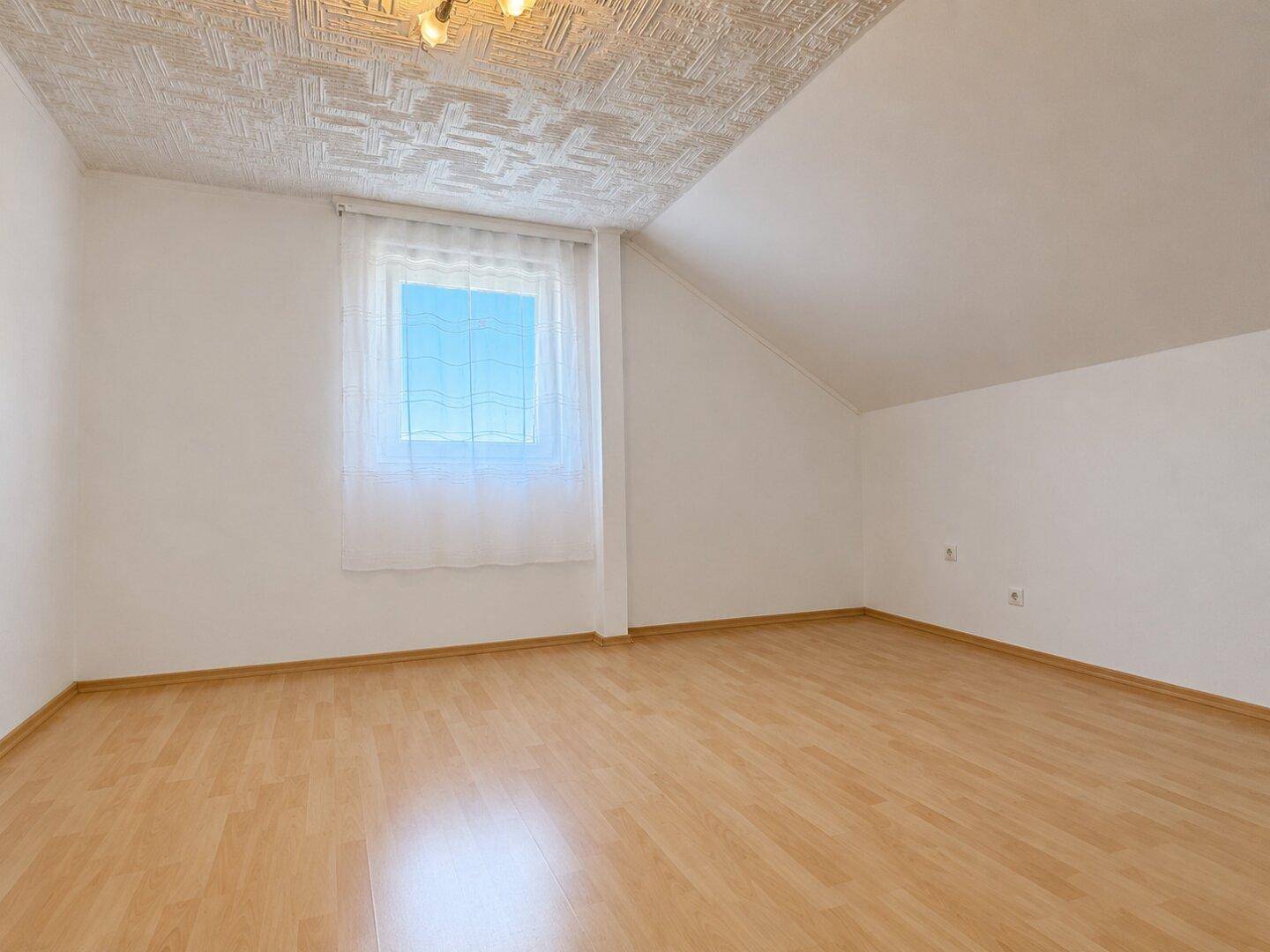 Geräumiges Zimmer im Dachgeschoss mit schräger Decke, Laminatboden und Fenster.