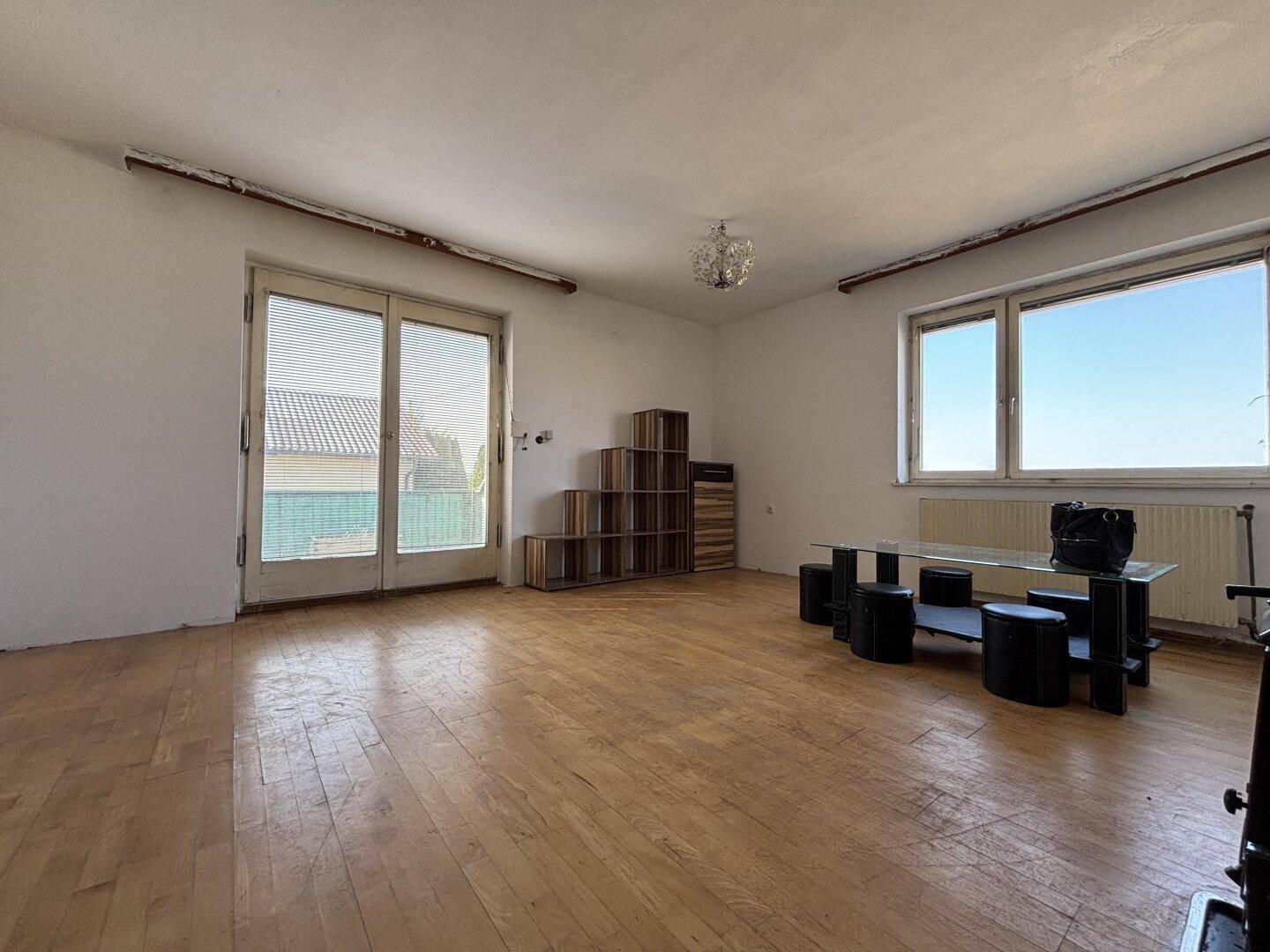 Geräumiges Wohnzimmer mit Holzparkett, großen Fenstern und Zugang zu einem Außenbereich.