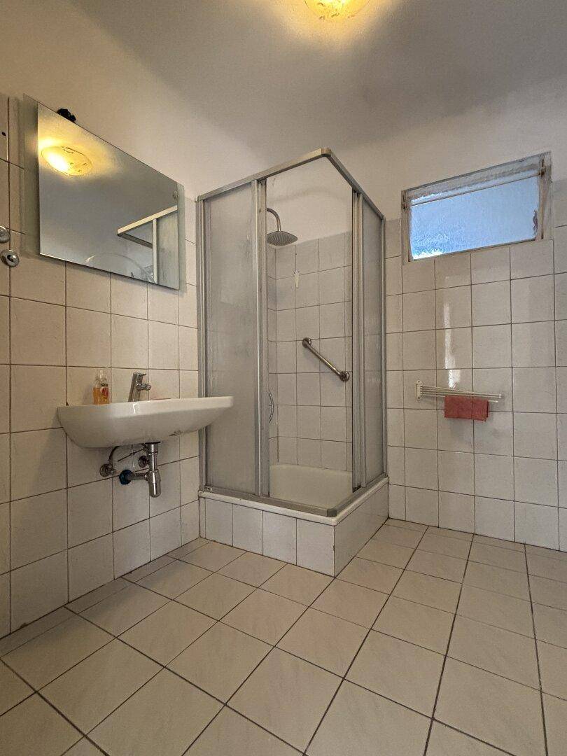 Gefliestes Badezimmer mit Duschkabine, Waschbecken und kleinem Fenster für Belüftung.