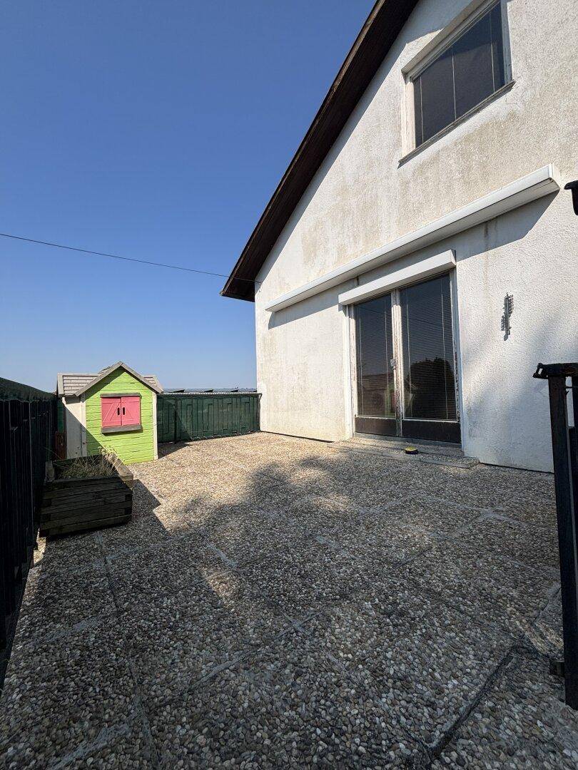 Gepflasterte Terrasse hinter dem Haus mit einem kleinen bunten Gartenhaus unter blauem Himmel.