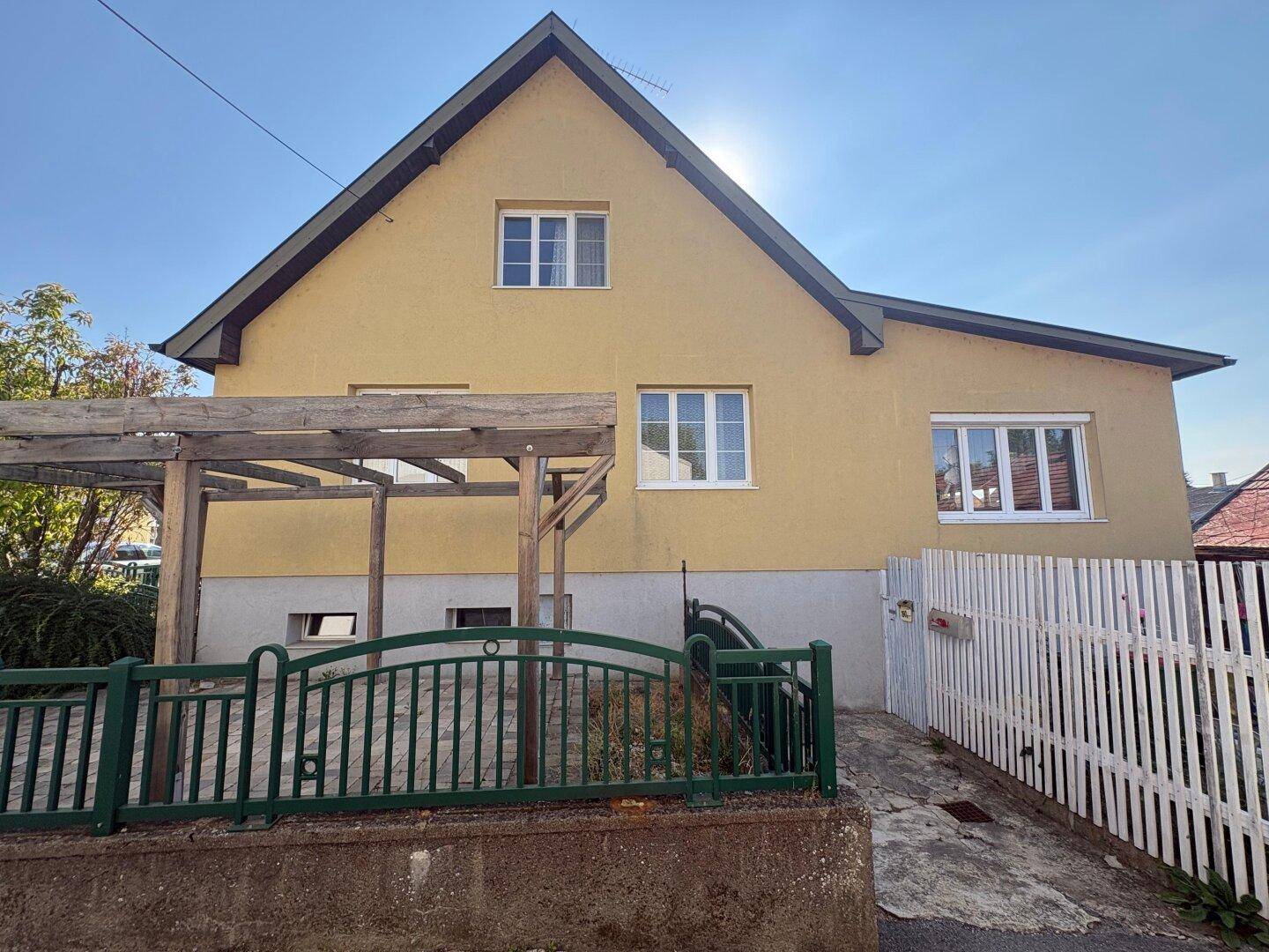 Seitenansicht eines gelben Hauses mit Satteldach und einer Holzpergola im Vorgartenbereich.