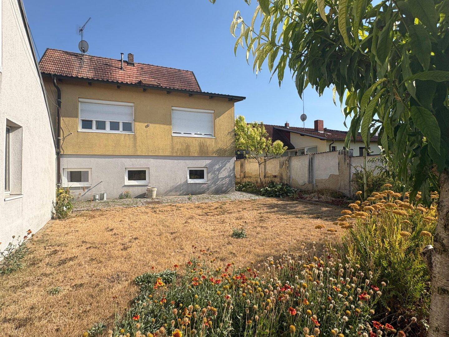 Rückansicht des Hauses mit einem trockenen Rasen und blühenden Blumen im Gartenbereich.