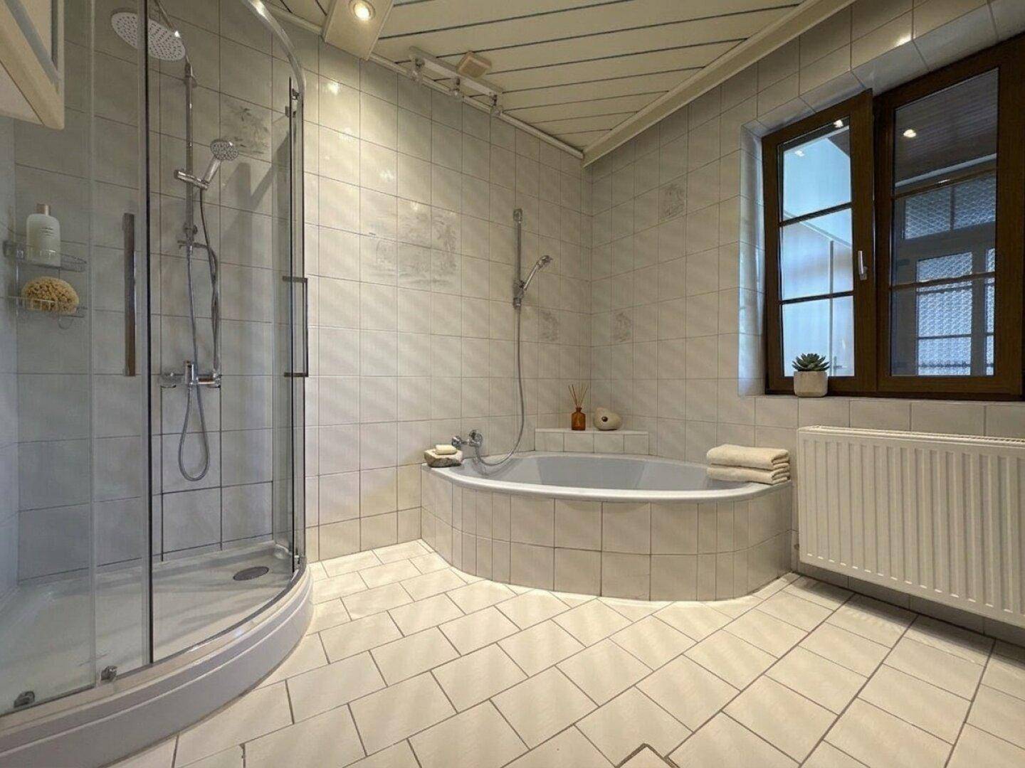 Badezimmer mit weißen Fliesen, einer Duschkabine und einer runden Badewanne unter einem Fenster.