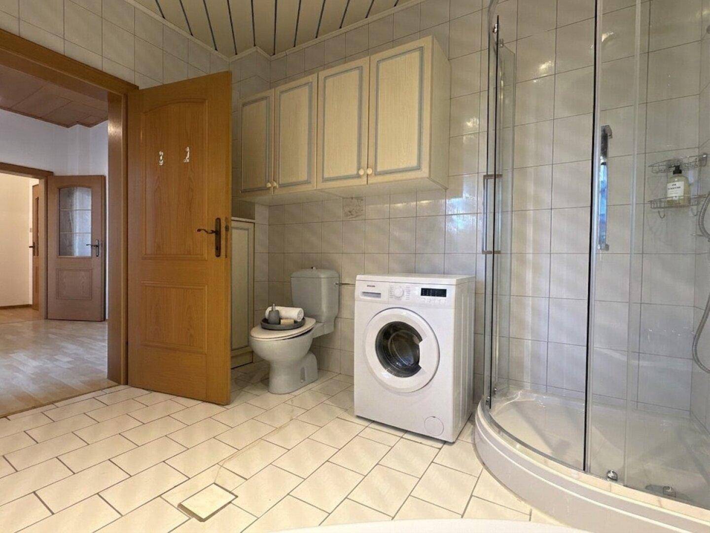 Badezimmer mit Waschmaschine, Toilette und Duschkabine, alles in weißen Fliesen gehalten.