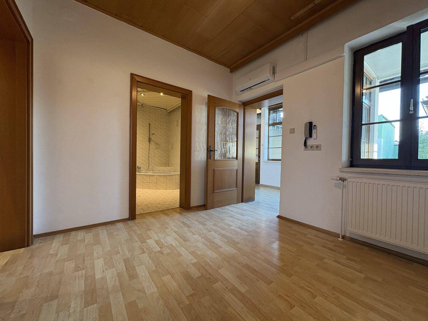 Heller Raum mit Holzboden, Klimaanlage und Zugang zu einem Badezimmer und weiteren Räumen.