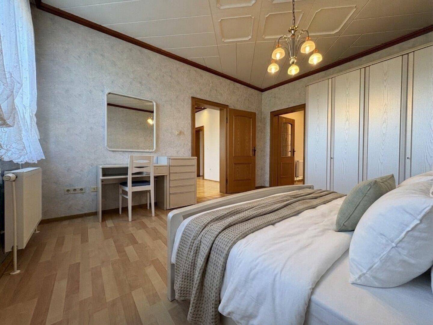 Schlafzimmer mit Holzboden, weißem Kleiderschrank und einem Schminktisch neben dem Bett.