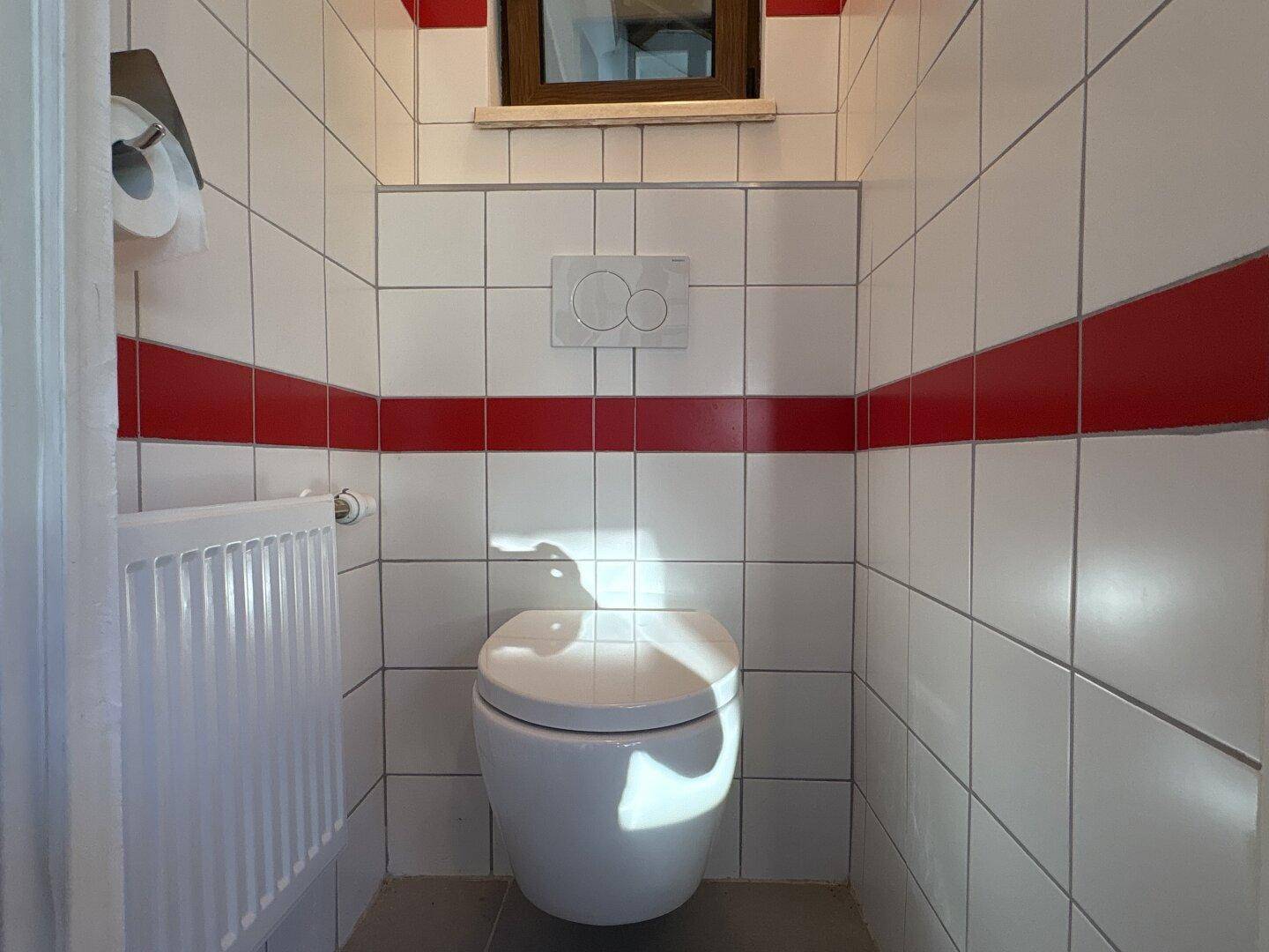 Kleines WC mit weißen Fliesen und einem roten Streifen, ausgestattet mit einer Toilette.