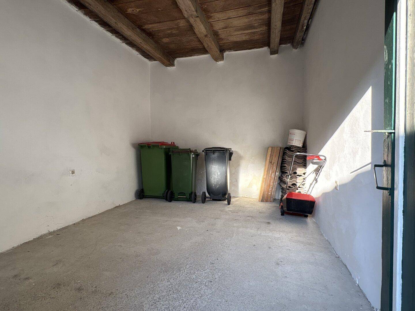 Einfacher Abstellraum mit Betonboden und sichtbaren Holzbalken an der Decke.