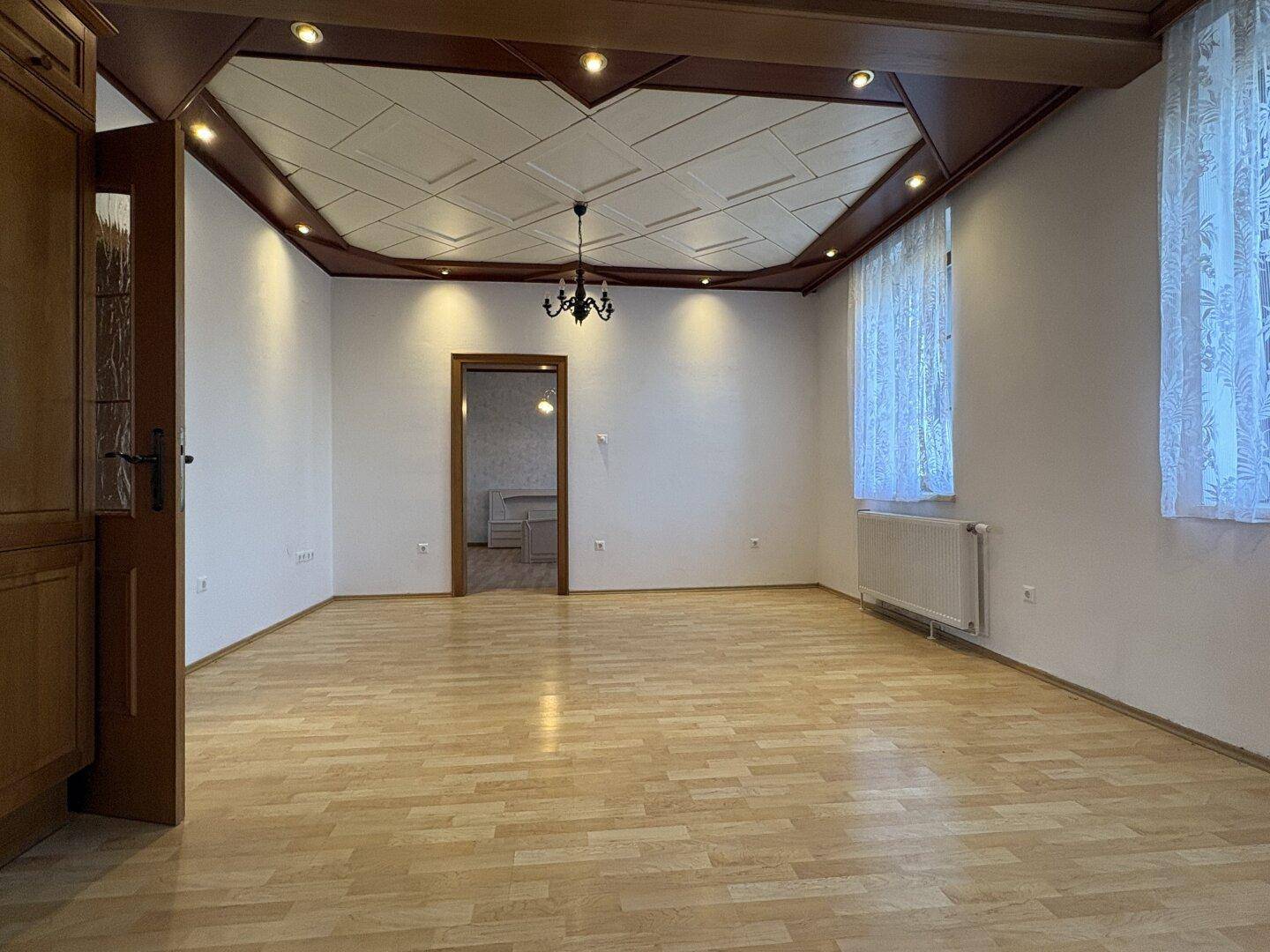 Heller Wohnbereich mit Holzboden, dekorativer Holzdecke und Fenstern mit Vorhängen.