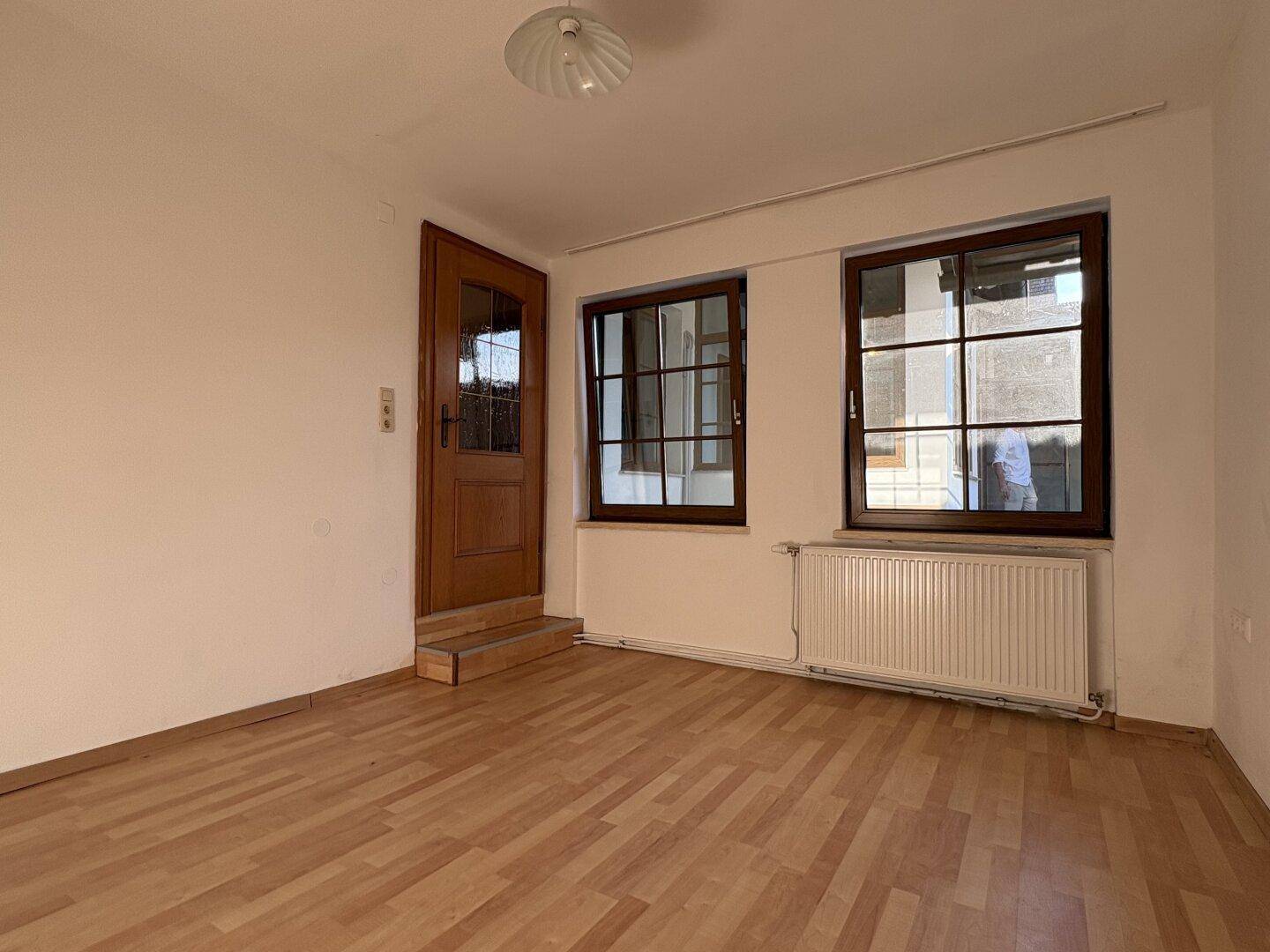 Heller Raum mit Holzboden, zwei Fenstern und einer Holztür, die nach draußen führt.