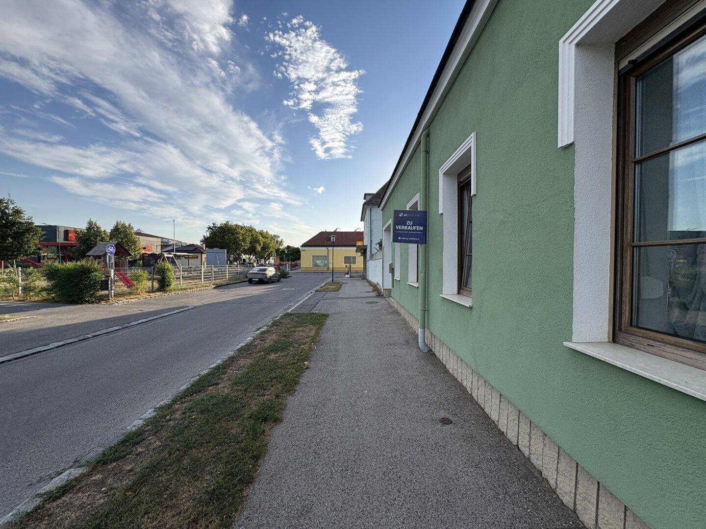 Grüne Hausfassade mit Fenstern und einem "Verkaufen"-Schild an einer ruhigen Straße.