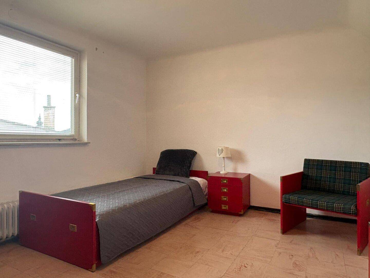 Schlafzimmer mit Einzelbett, rotem Nachttisch und kariertem Sessel.