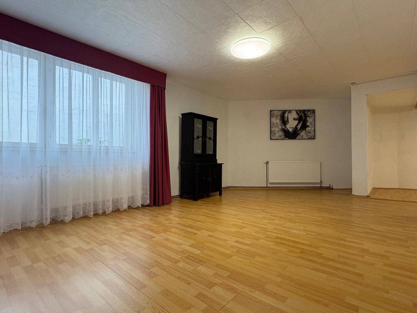 Großes, helles Wohnzimmer mit Laminatboden und rotem Vorhang.
