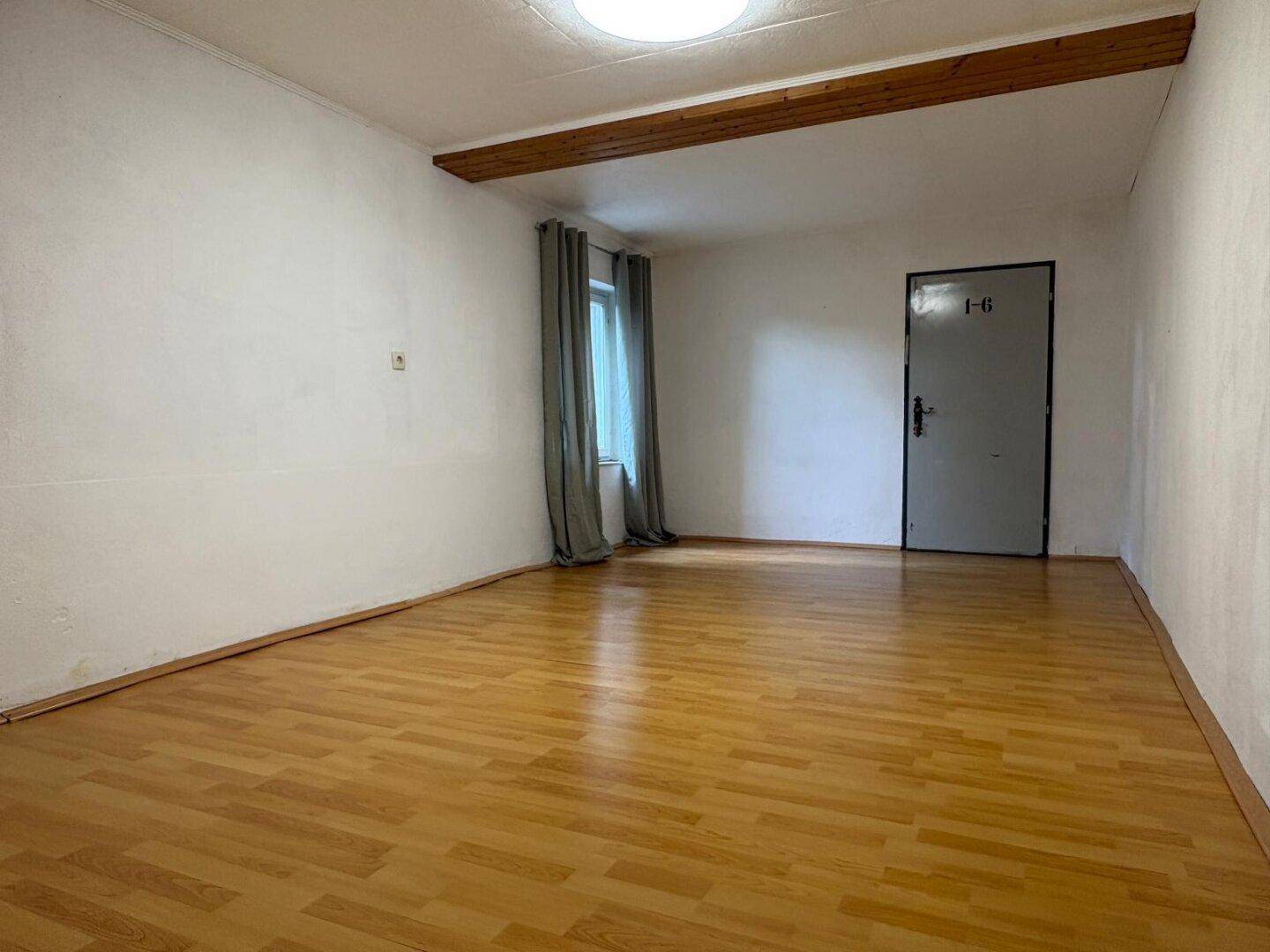 Heller Raum mit Laminatboden, Holzbalken und Fenster mit Vorhang.