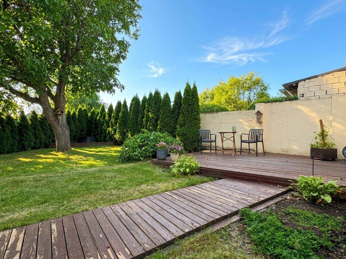 Sonniger Garten mit Holzterrasse, Sitzbereich und hohen Hecken für Privatsphäre.