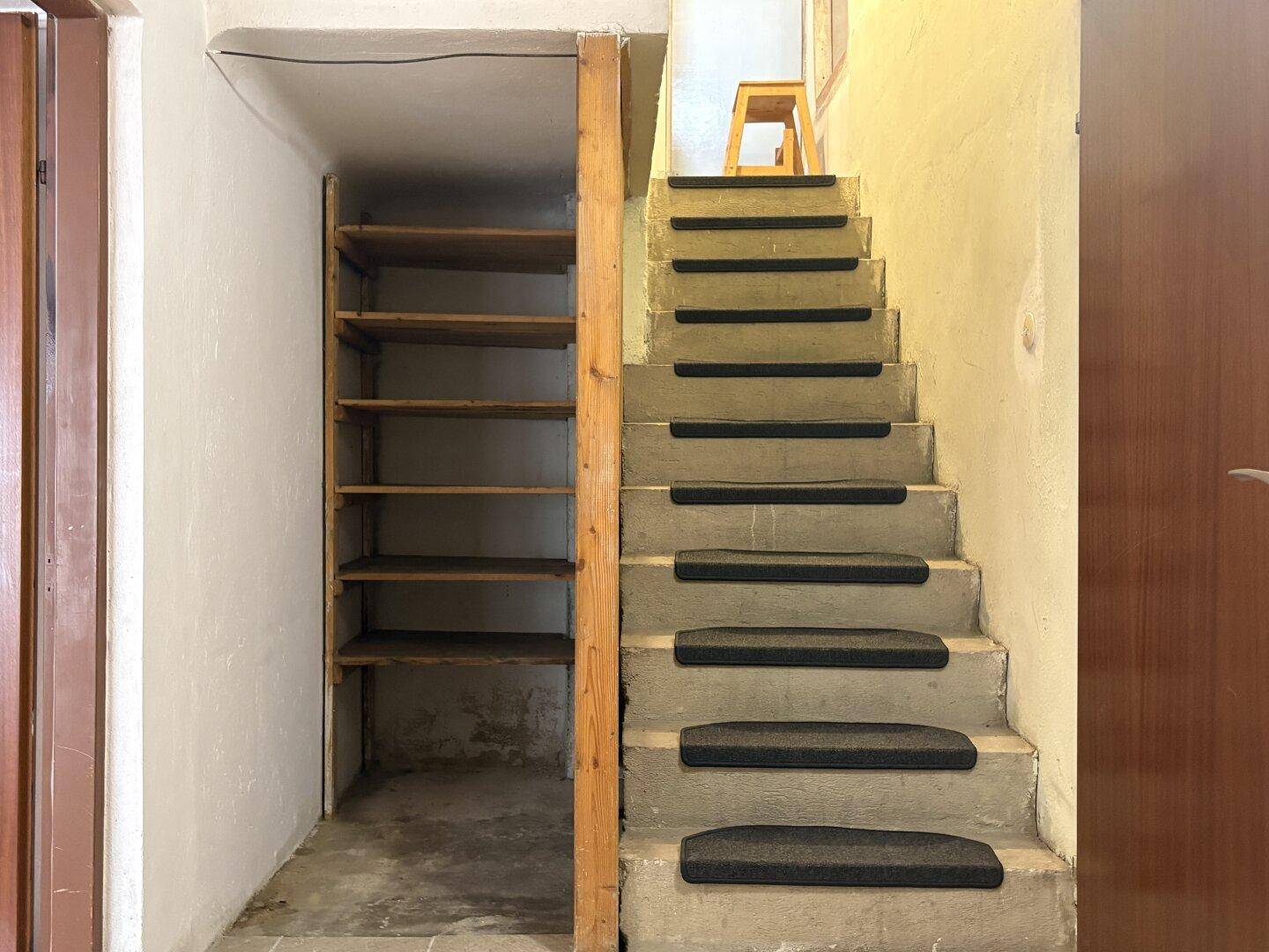 Kellerraum mit Betontreppe, Holzregalen und einfacher Beleuchtung.