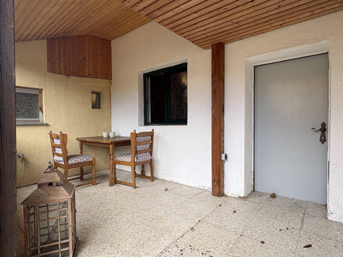 Überdachter Außenbereich mit kleinem Holztisch, zwei Stühlen und Fliesenboden, ideal zum Entspannen.