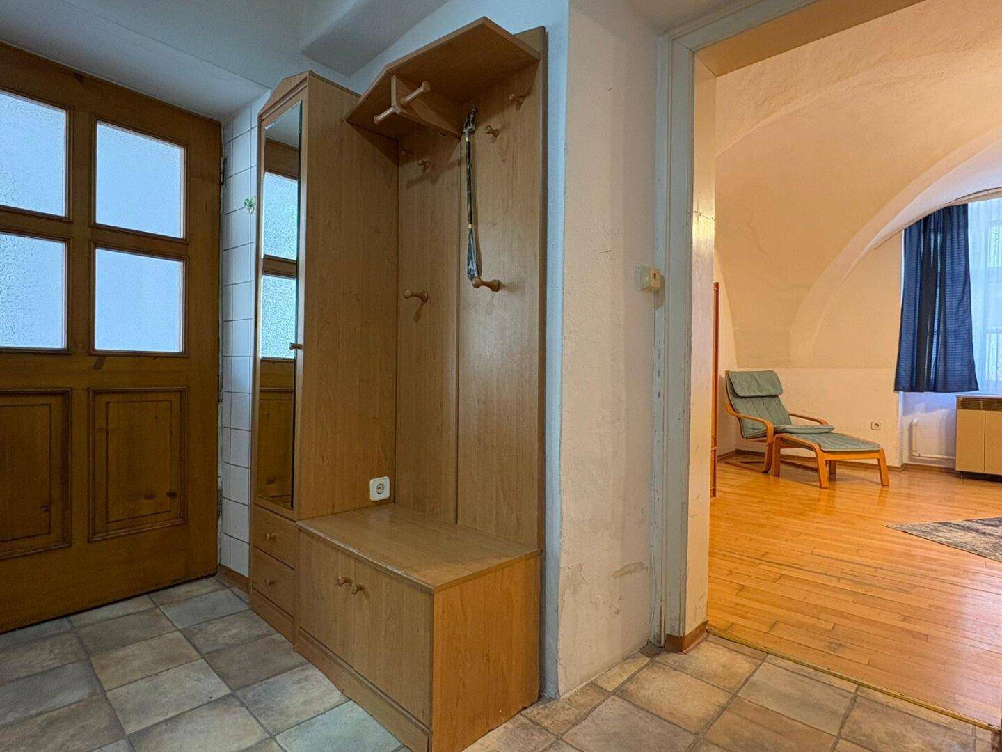 Eingangsbereich mit Holzgarderobe, Spiegel und gefliestem Boden, führt in einen Wohnraum.