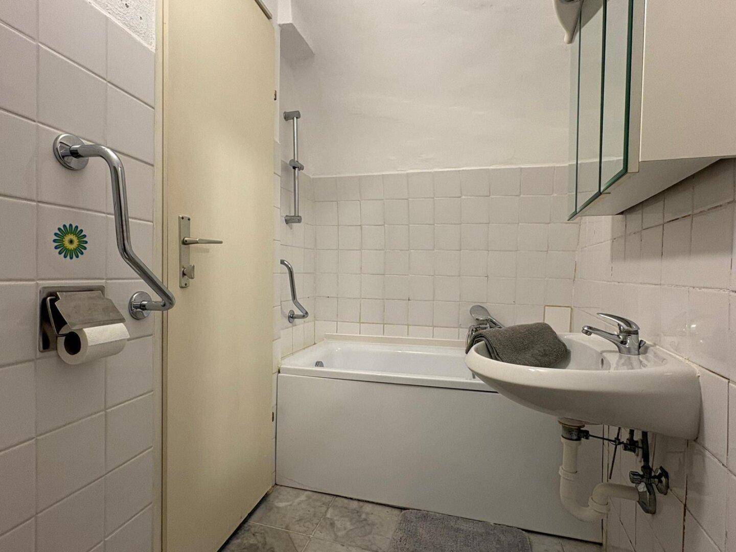 Helles Badezimmer mit Badewanne, Waschbecken und weißen Fliesen, ausgestattet mit Haltegriffen.