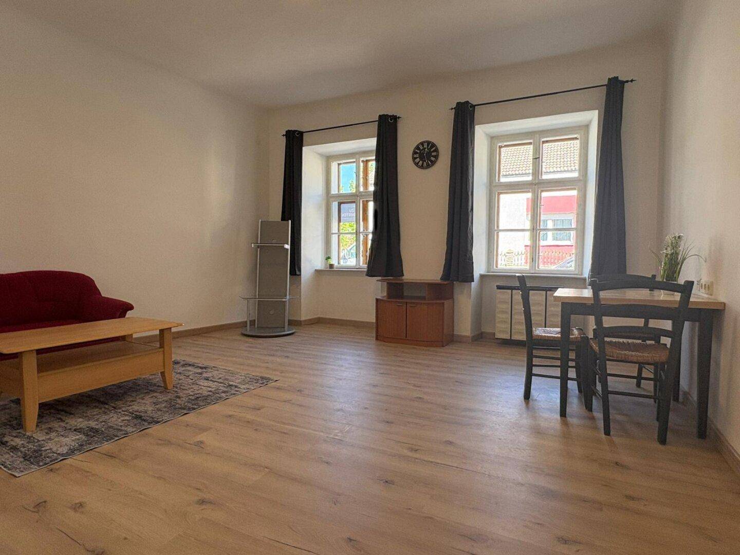 Heller Wohn-Essbereich mit Holzboden, Esstisch und großen Fenstern mit dunklen Vorhängen.