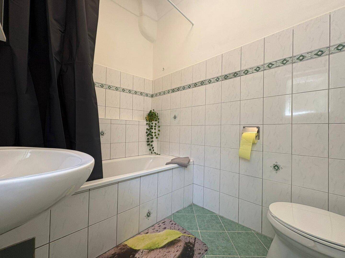 Badezimmer mit Badewanne, WC und grün-weißen Fliesen, ausgestattet mit Duschvorhang.