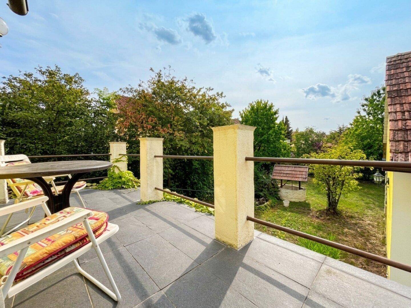 Sonniger Balkon mit Sitzgelegenheiten und Blick auf den grünen Garten mit Brunnen.
