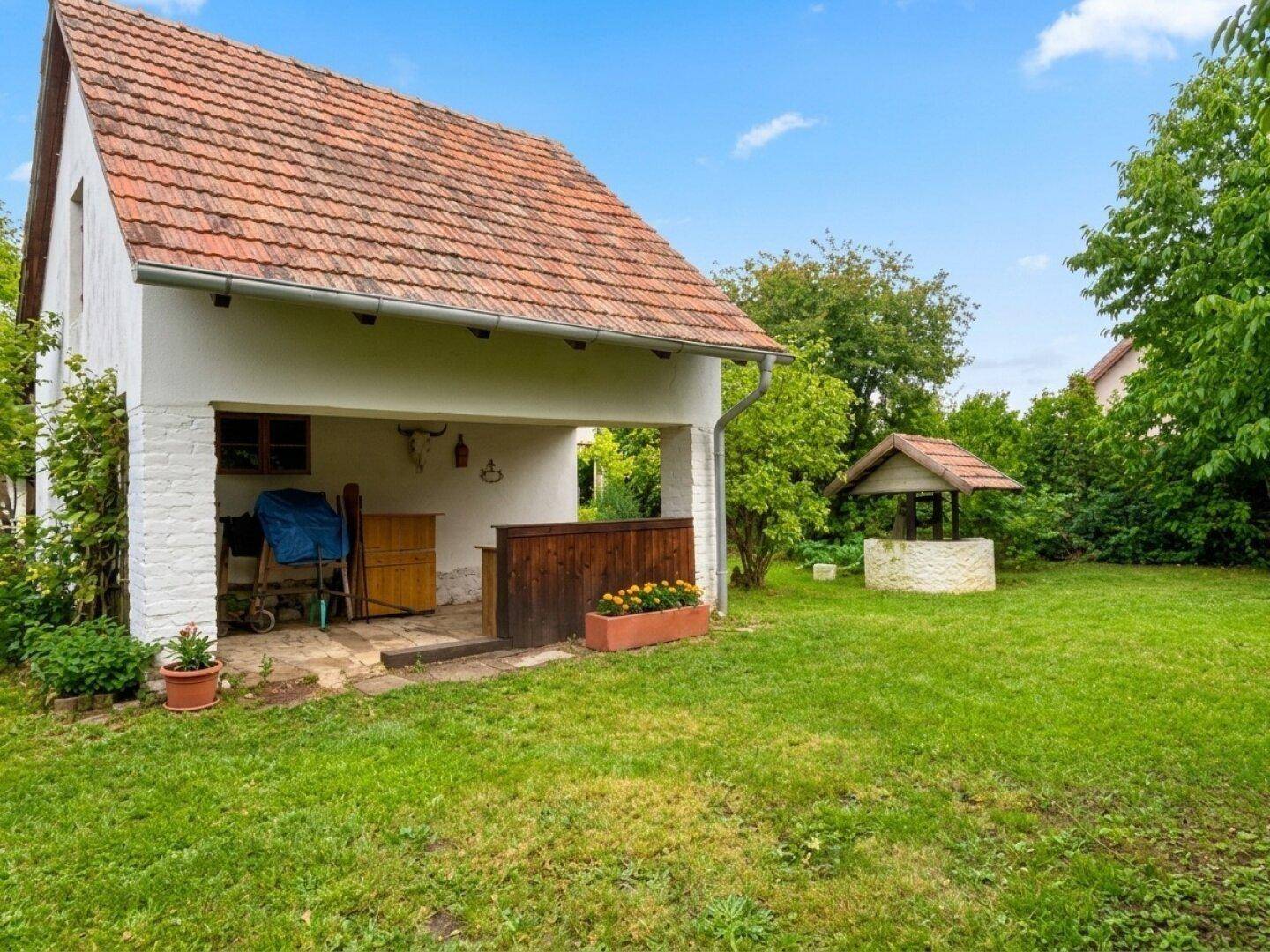 Seitlicher Blick auf das Haus mit überdachtem Bereich und dem weitläufigen Garten mit Brunnen.