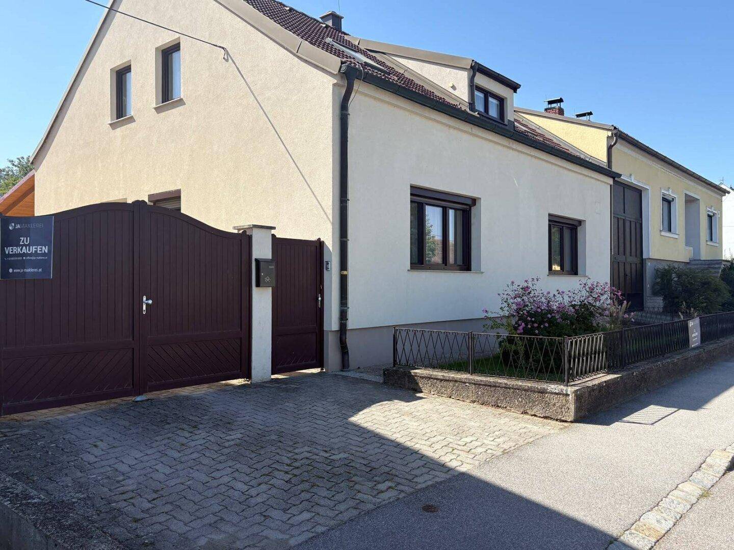 Vorderansicht eines gepflegten Hauses mit beiger Fassade, braunem Tor und einem kleinen Vorgarten.