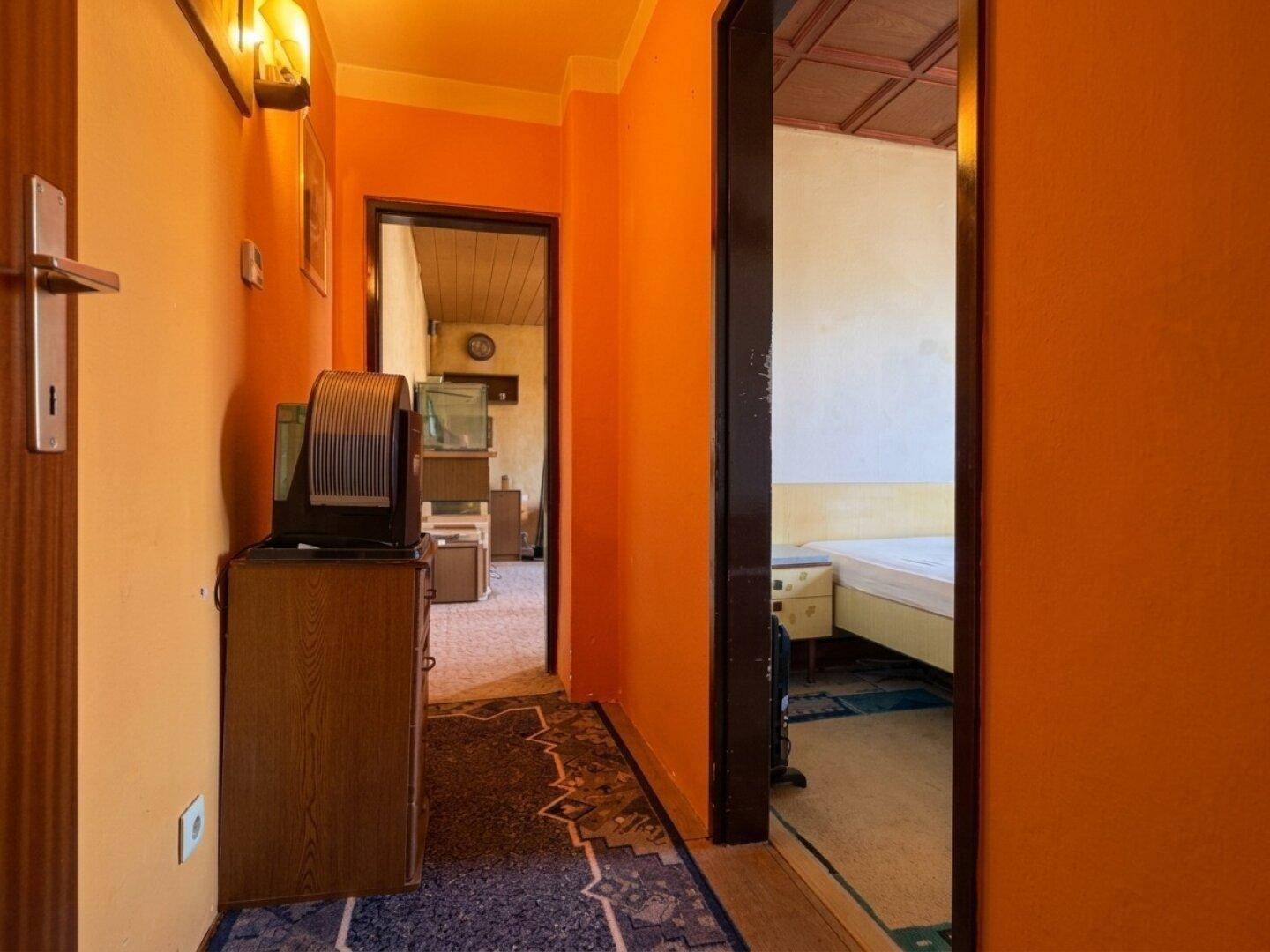 Orangefarbener Flur mit Zugang zu einem Schlafzimmer und einem weiteren Raum, ausgestattet mit älteren Möbeln.