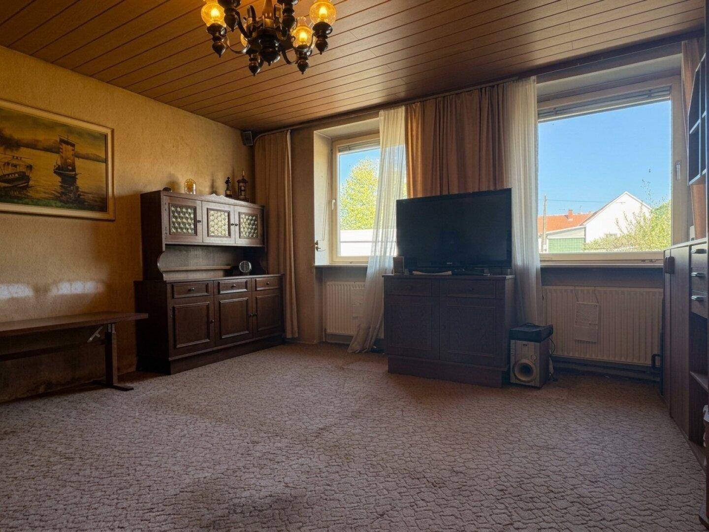 Wohnzimmer mit Teppichboden, Holzdecke und zwei Fenstern, eingerichtet mit traditionellen Holzmöbeln.