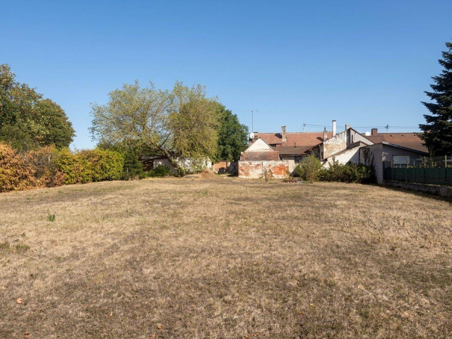 Großes, trockenes Grasfeld mit älteren Gebäuden im Hintergrund unter blauem Himmel.