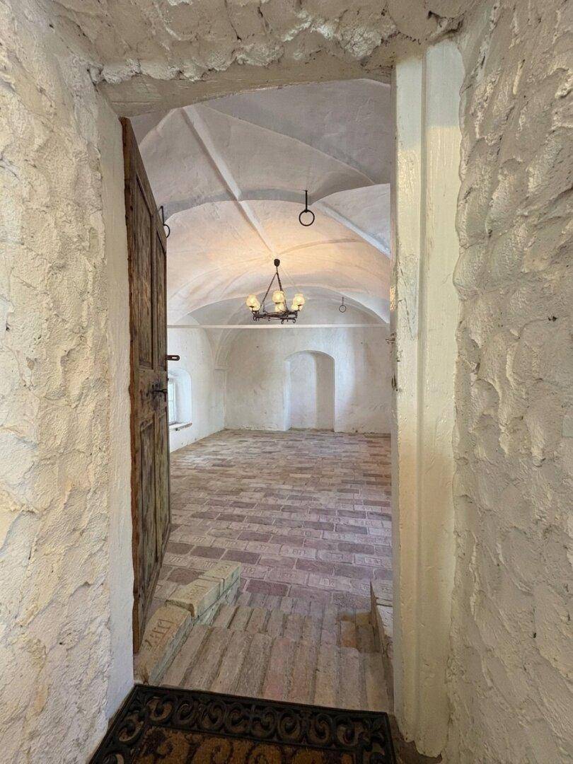 Blick durch eine rustikale Holztür in einen historischen Raum mit Gewölbedecke und Ziegelboden.