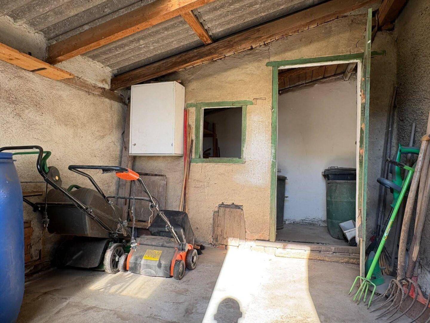 Rustikaler Abstellraum mit Gartengeräten, Rasenmäher und einem kleinen Fenster.