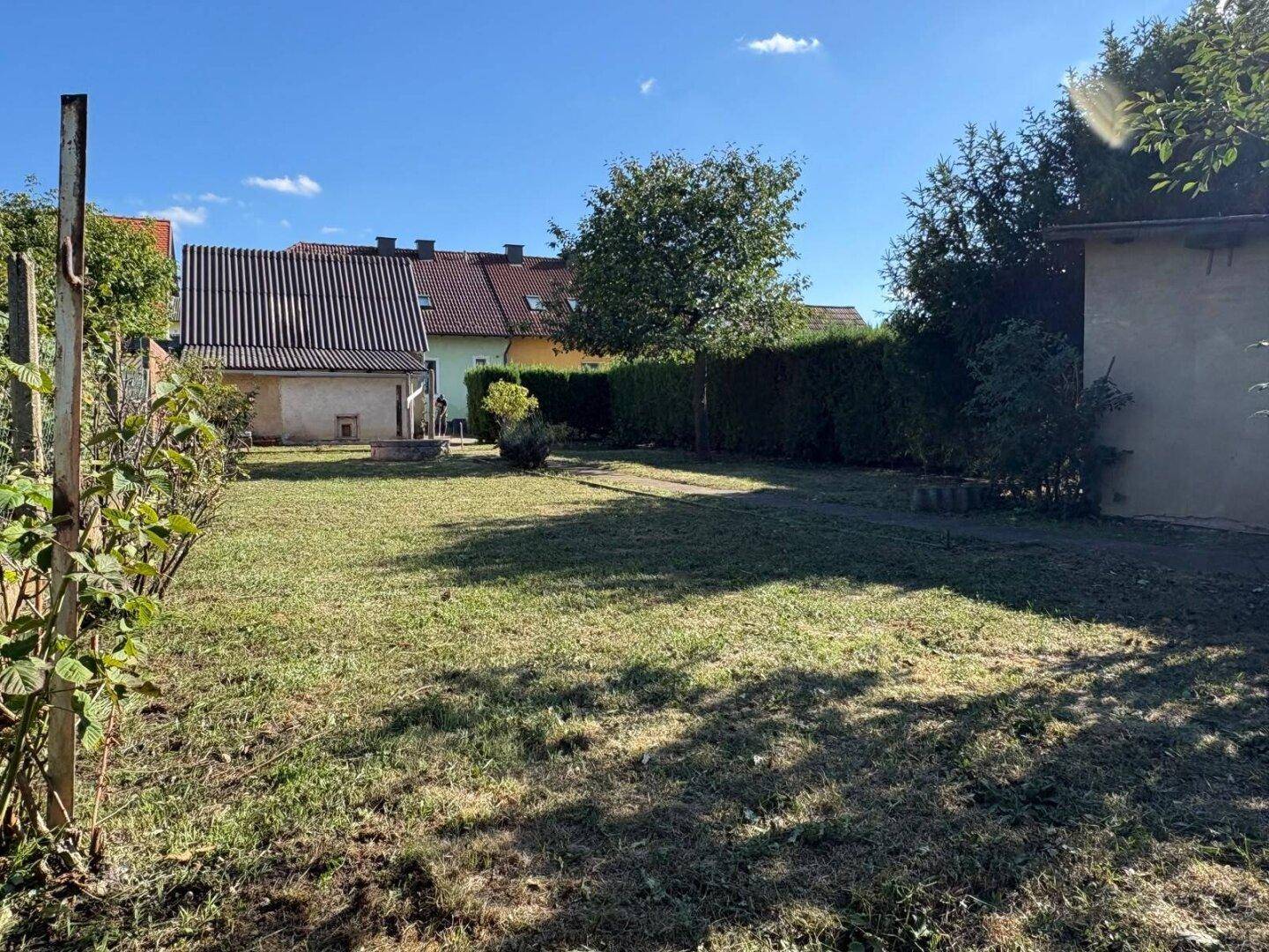 Weitläufiger Gartenbereich mit Rasen, Hecke und Blick auf Nachbargebäude, sonnig und grün.