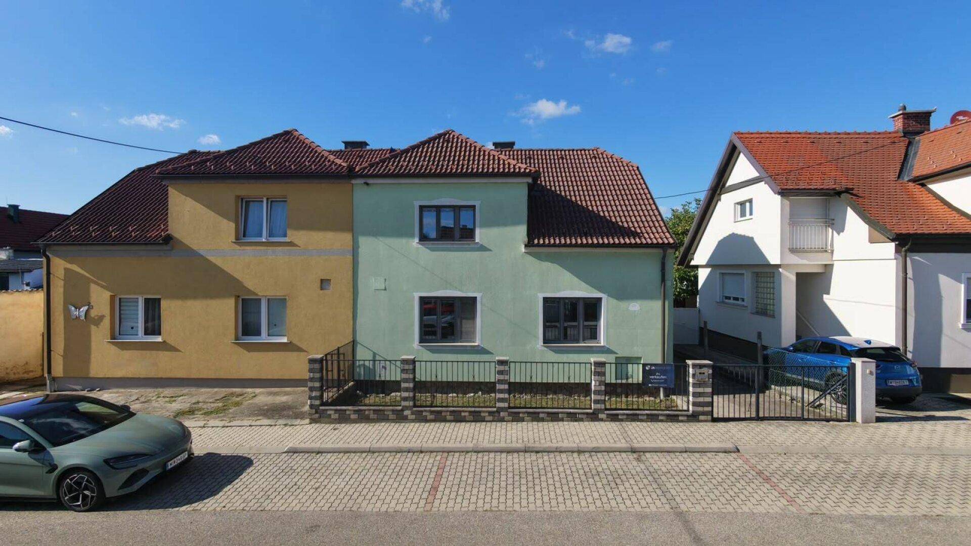 Ansicht von zwei benachbarten Häusern mit unterschiedlichen Fassadenfarben und Ziegeldächern an einem sonnigen Tag.