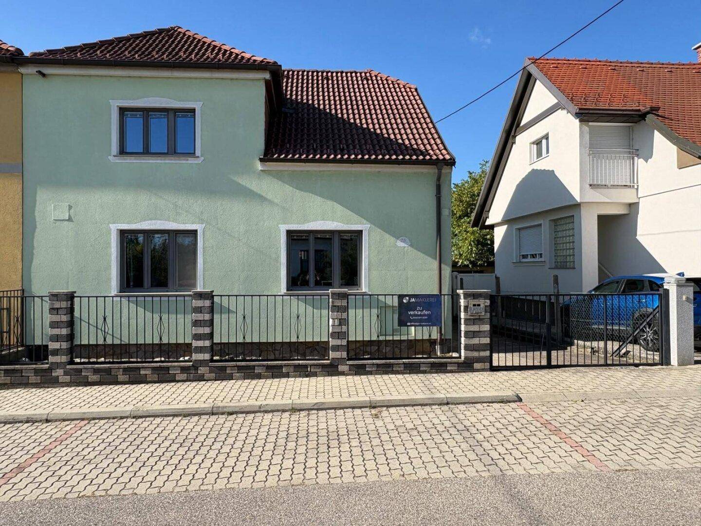 Vorderansicht eines Einfamilienhauses mit hellgrüner Fassade und Ziegeldach, umgeben von einem Zaun.