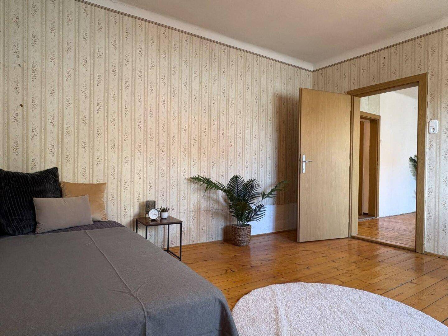 Schlafzimmer mit Holzdielenboden, Bett und Zimmerpflanze, Zugang zu weiterem Raum.