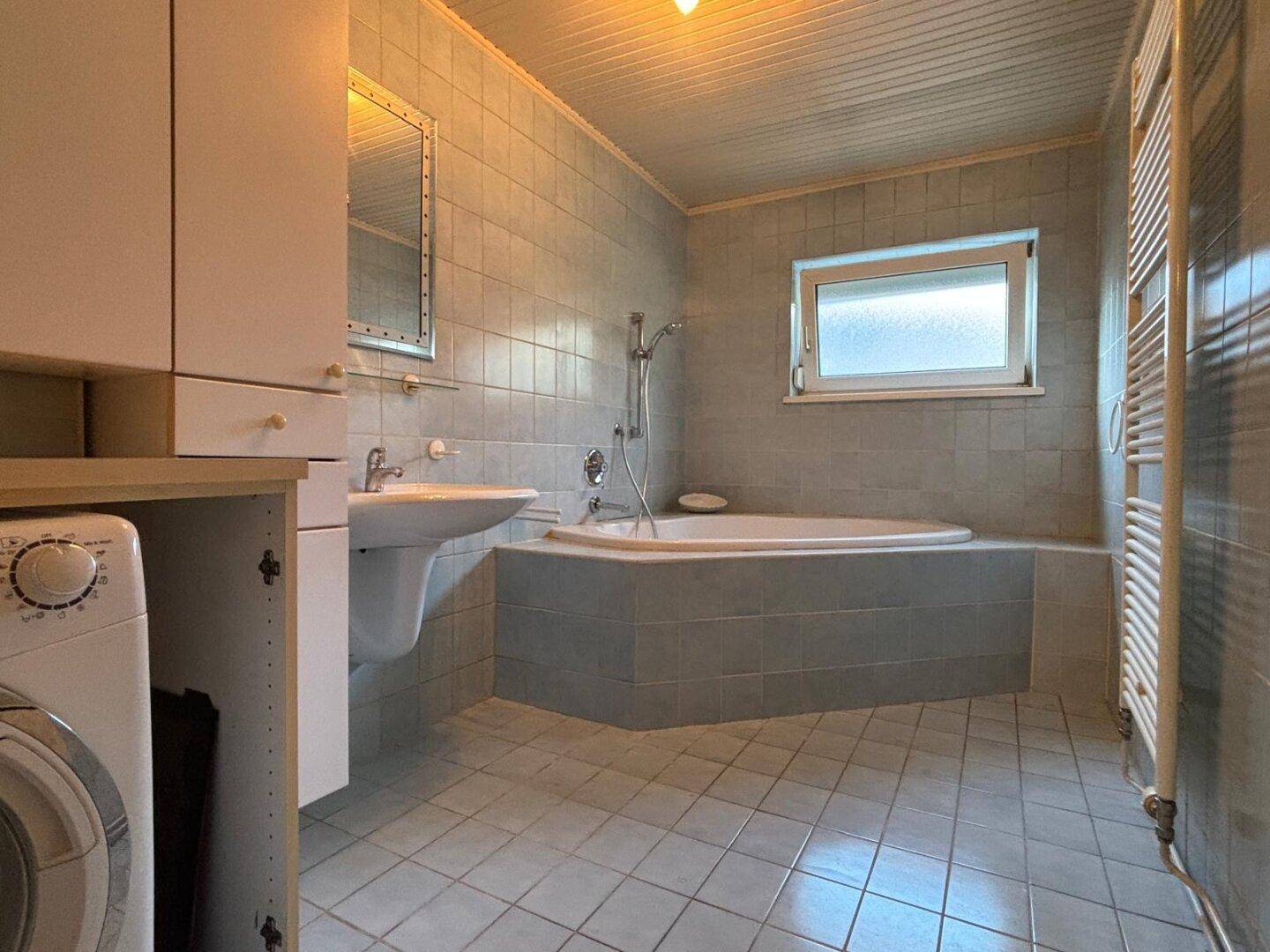 Gefliestes Badezimmer mit Badewanne, Waschbecken und Fenster für natürliche Belüftung.