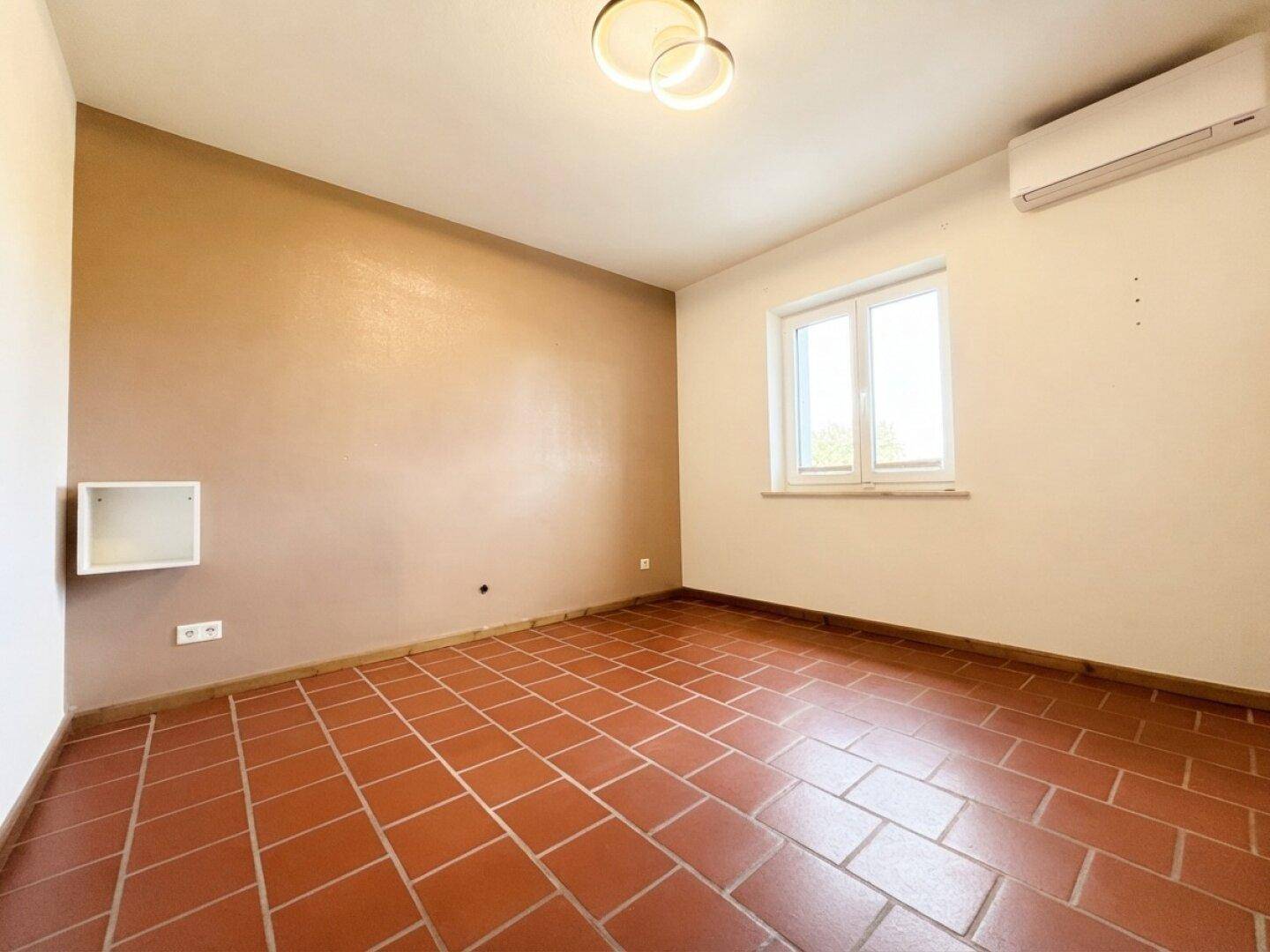 Helles Zimmer mit Terrakottafliesen, Klimaanlage und Fenster, bereit zur individuellen Gestaltung.