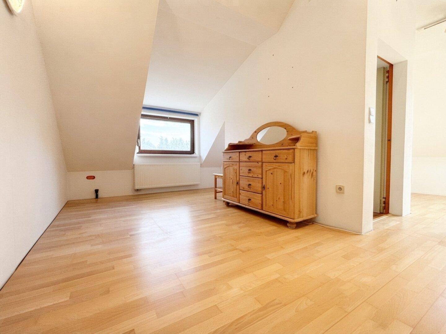 Geräumiger Dachbodenraum mit Holzboden, Fenster und rustikaler Kommode, ideal für ein Schlafzimmer.
