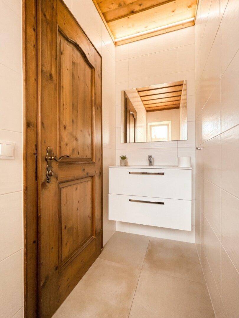 Modernes Badezimmer mit Waschtisch, Spiegel und Holztür, beleuchtet durch indirektes Licht.