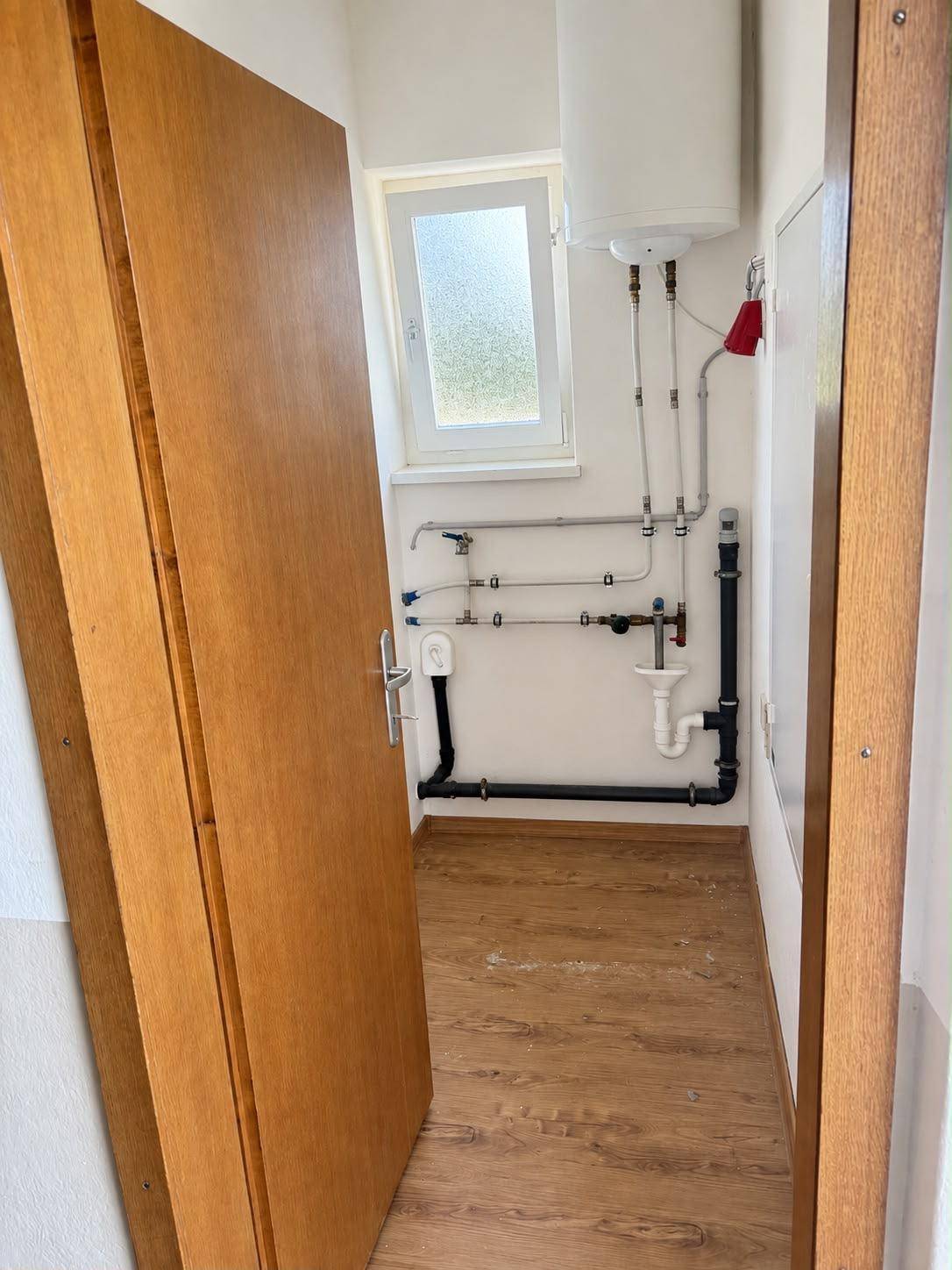 Kleiner Hauswirtschaftsraum mit Warmwasserboiler, sichtbaren Leitungen und Fenster.