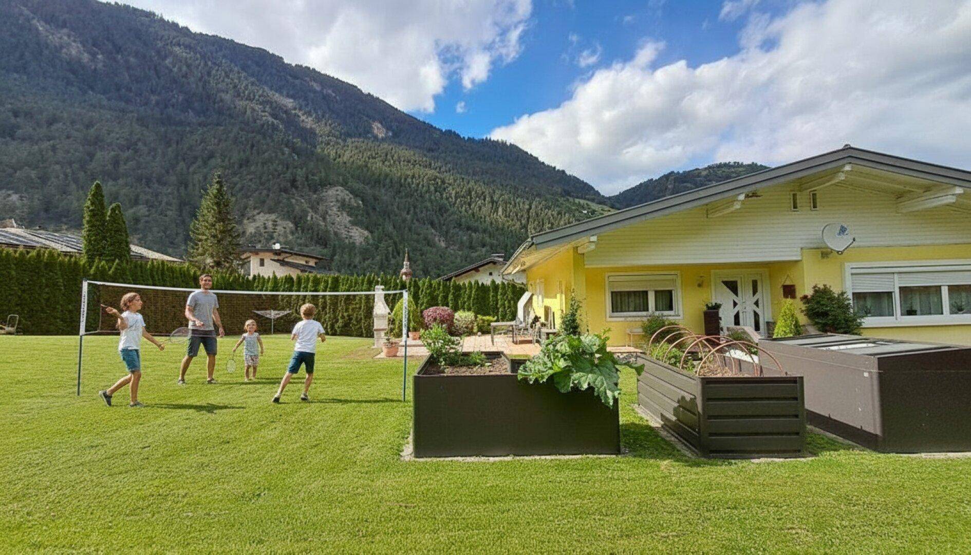 Familie spielt Badminton auf dem großen Rasen im Garten mit Hochbeeten und Bergblick.
