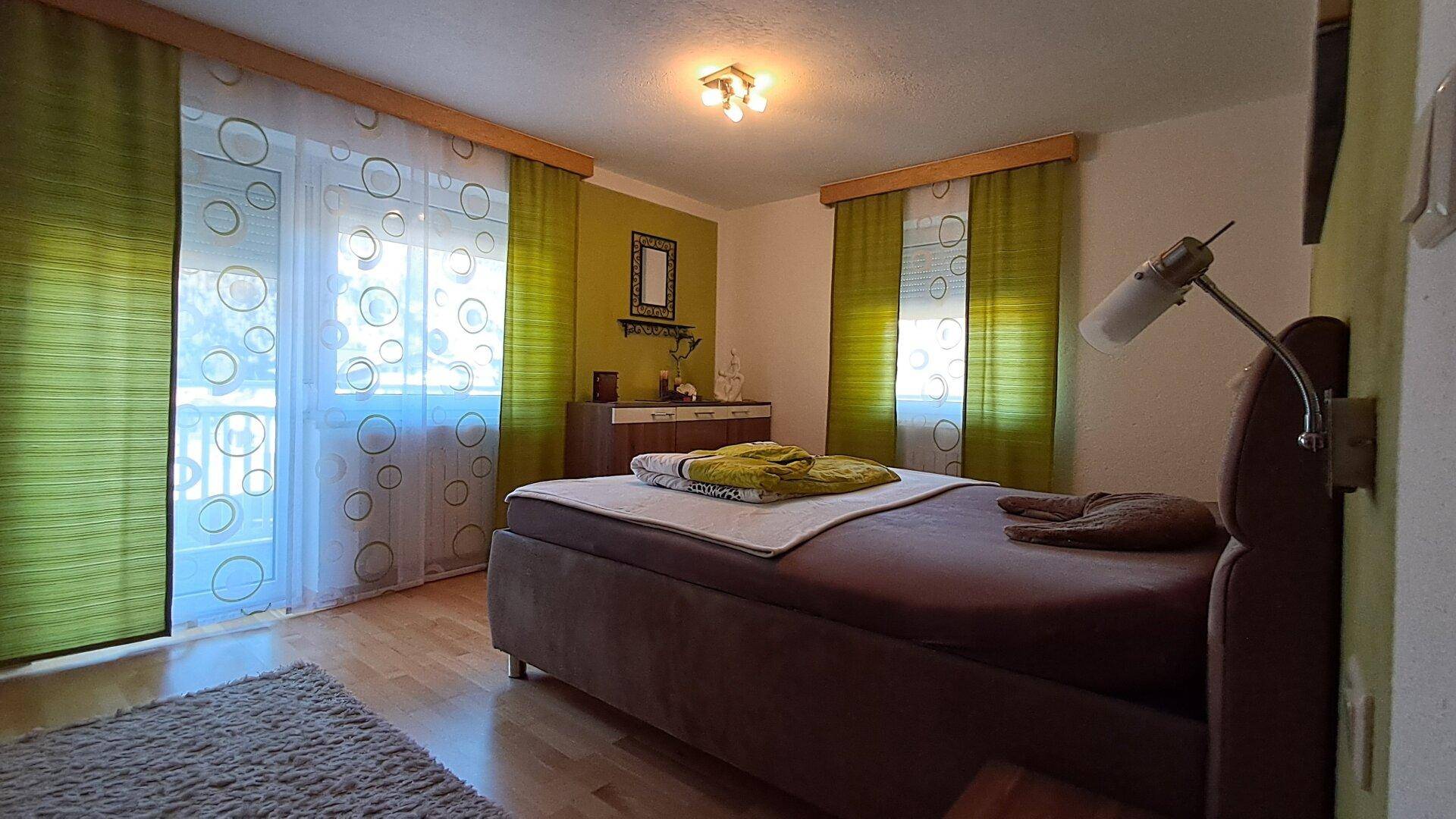 Helles Schlafzimmer mit Holzboden, grünen Vorhängen und Zugang zu einem Balkon.