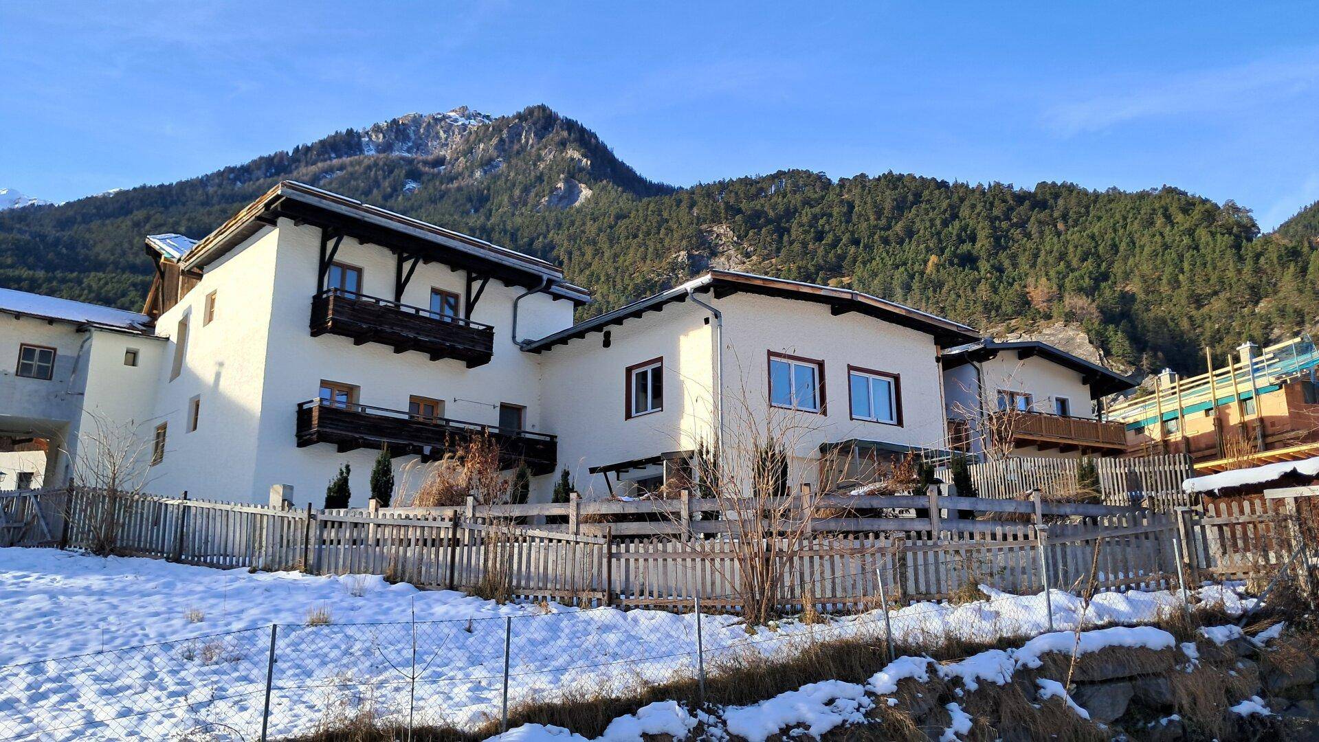 Mehrere Gebäude in einer verschneiten alpinen Landschaft mit Holzbalkonen und bewaldeten Bergen im Hintergrund.