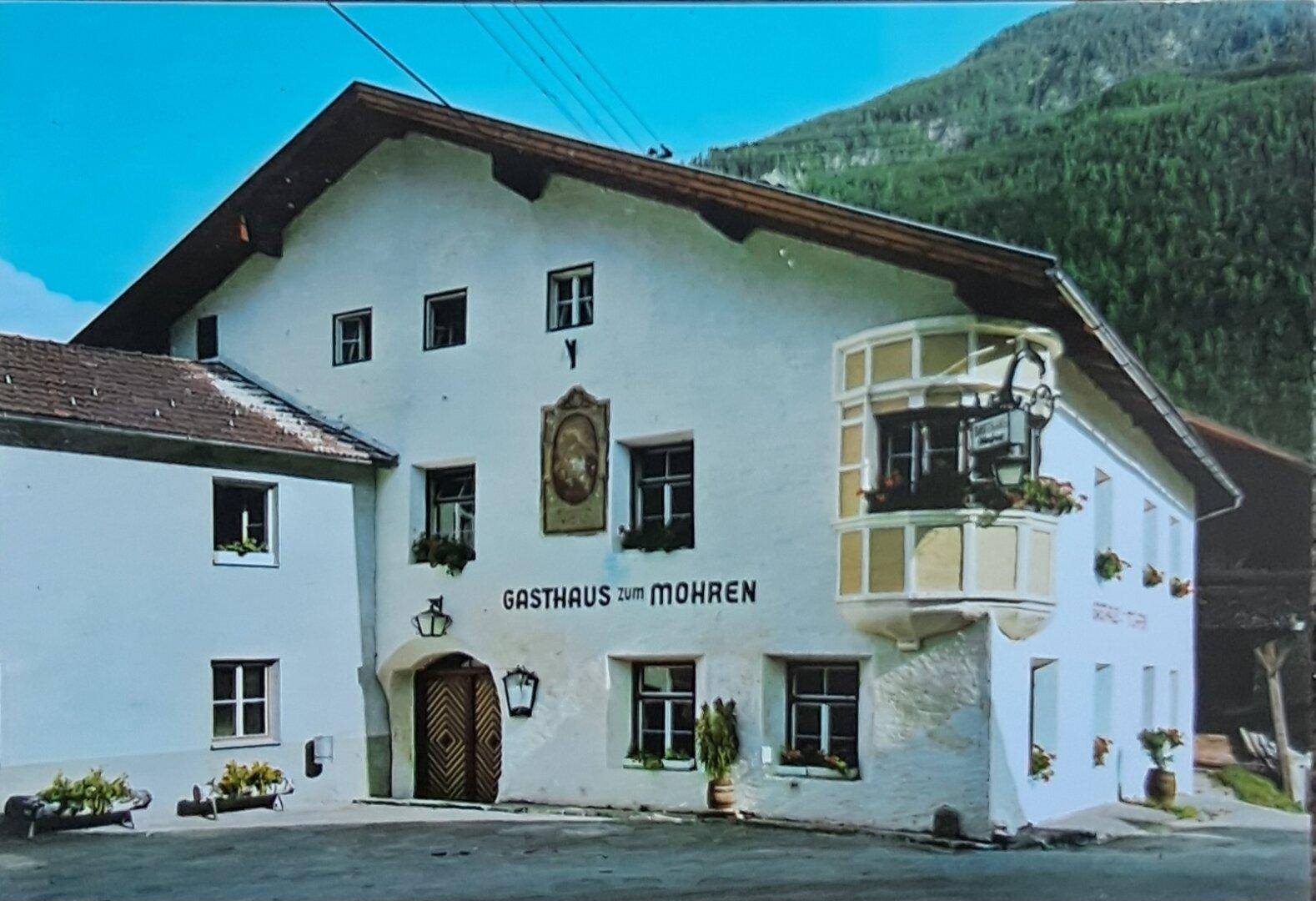 Historische Ansicht eines Gasthauses mit traditioneller Fassade, Erker und alpiner Bergkulisse im Hintergrund.