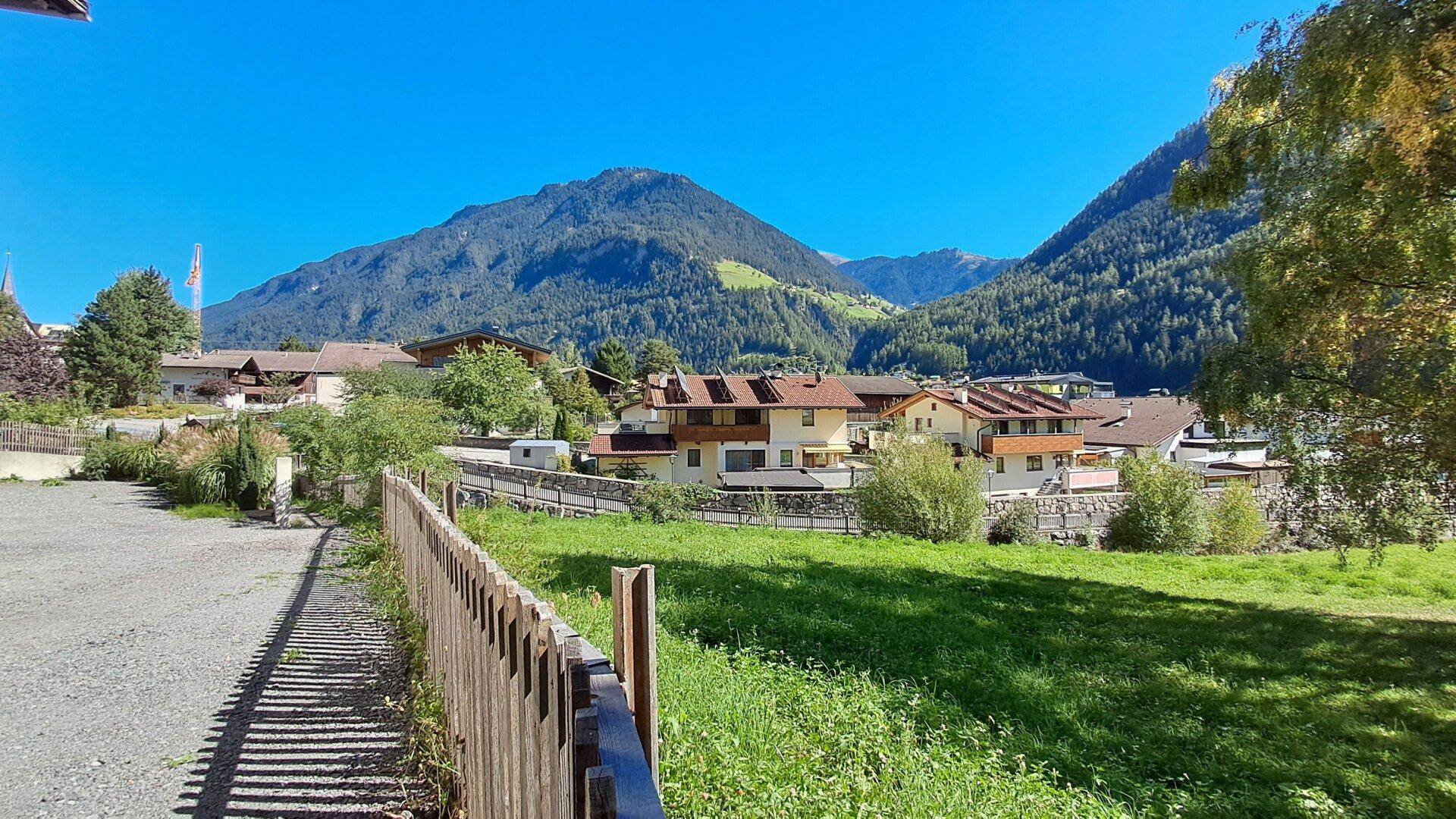 Panoramablick auf ein idyllisches Dorf mit grünen Wiesen und majestätischen Bergen unter blauem Himmel.