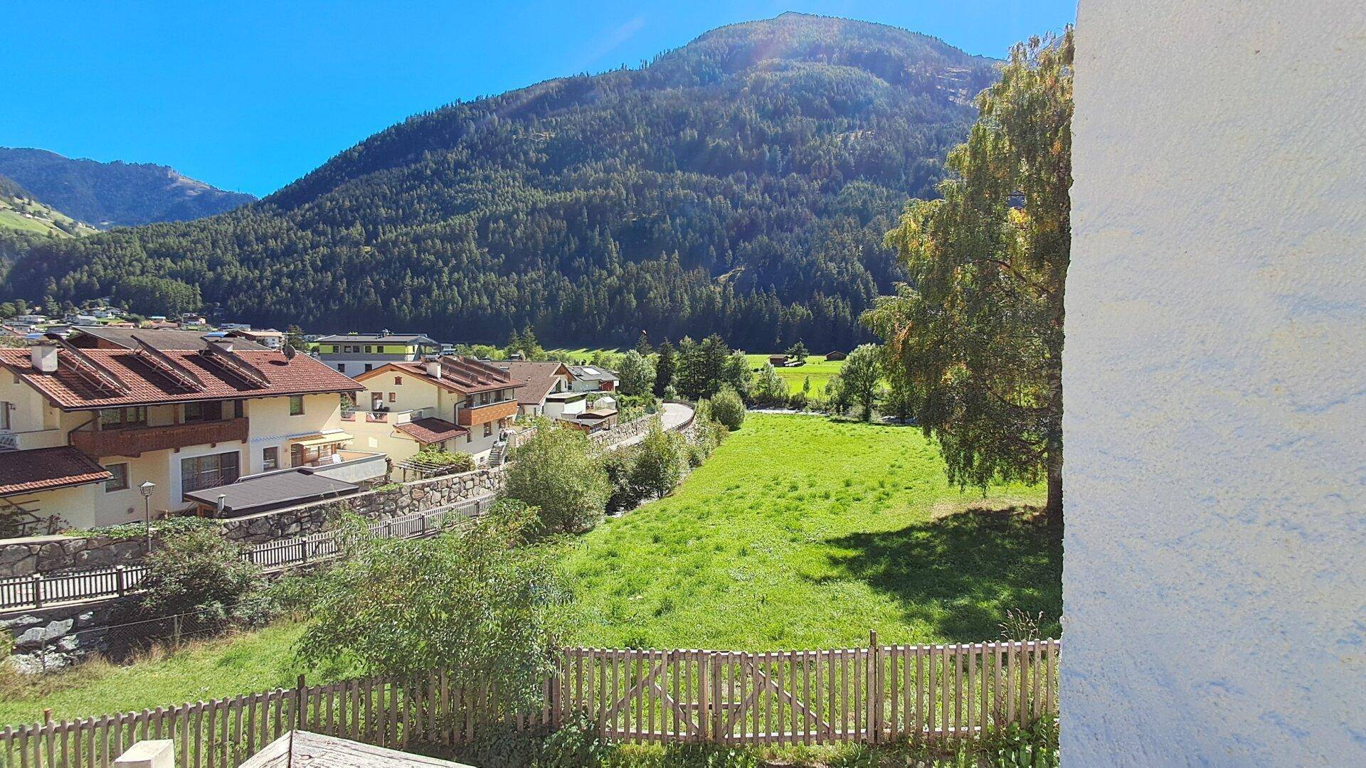 Aussicht auf ein Dorf mit grünen Feldern und bewaldeten Bergen, begrenzt durch einen Holzzaun im Vordergrund.